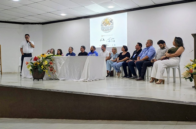 Pessoas sentadas em palco durante evento com apresentação e mesa principal