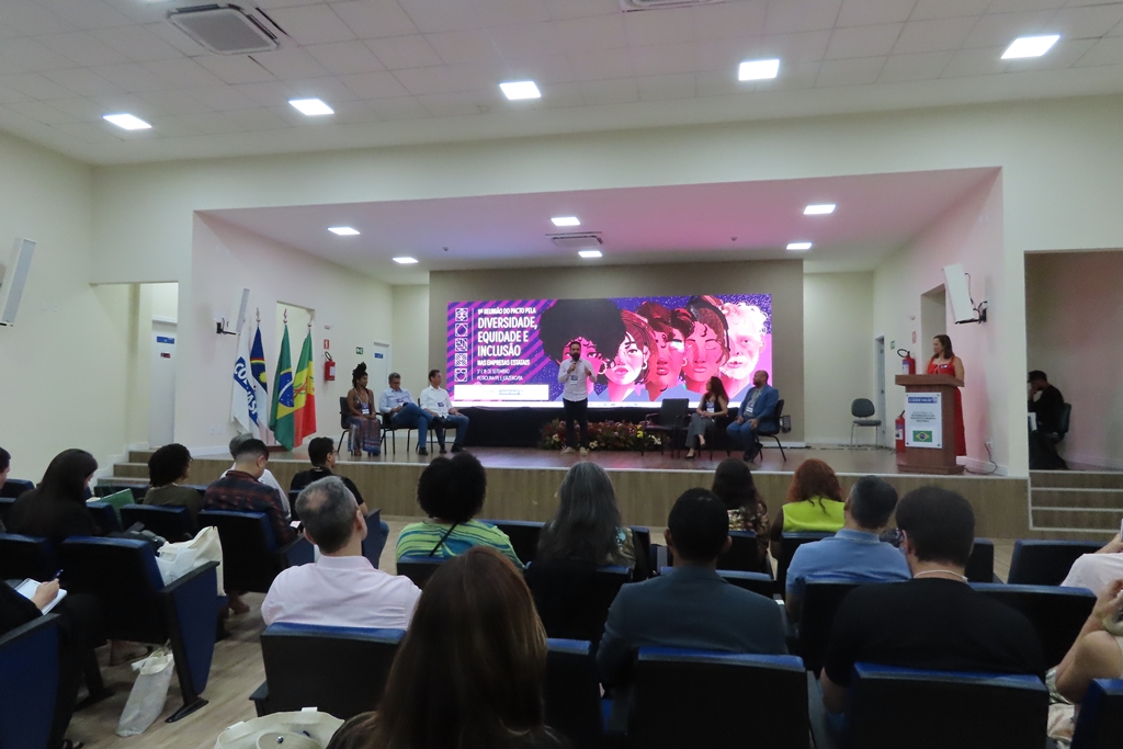 Palestrante fala ao público em auditório, com participantes no palco e painel sobre diversidade ao fundo.