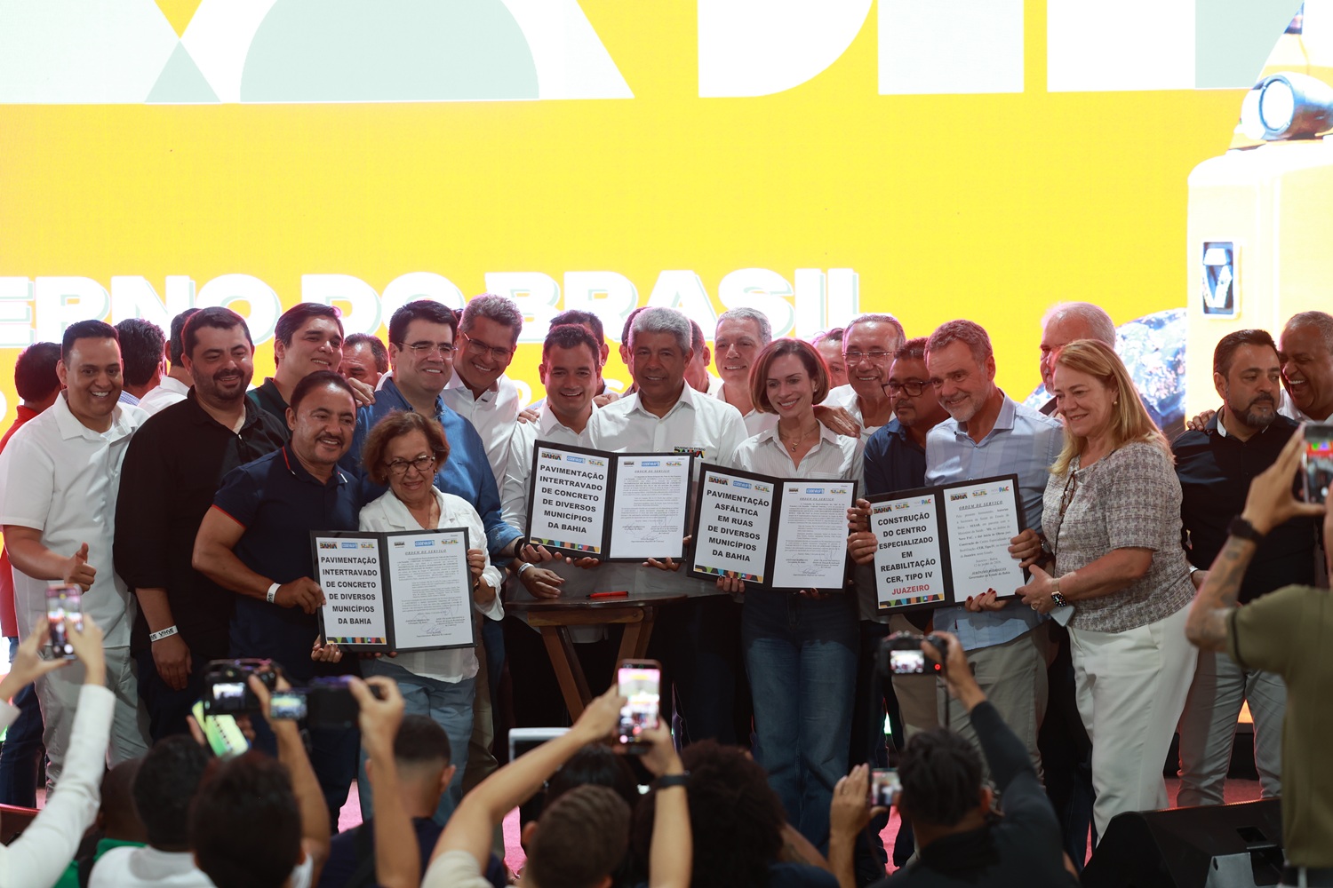 Autoridades no palco sorriem e seguram pastas de documentos oficiais abertas para o público