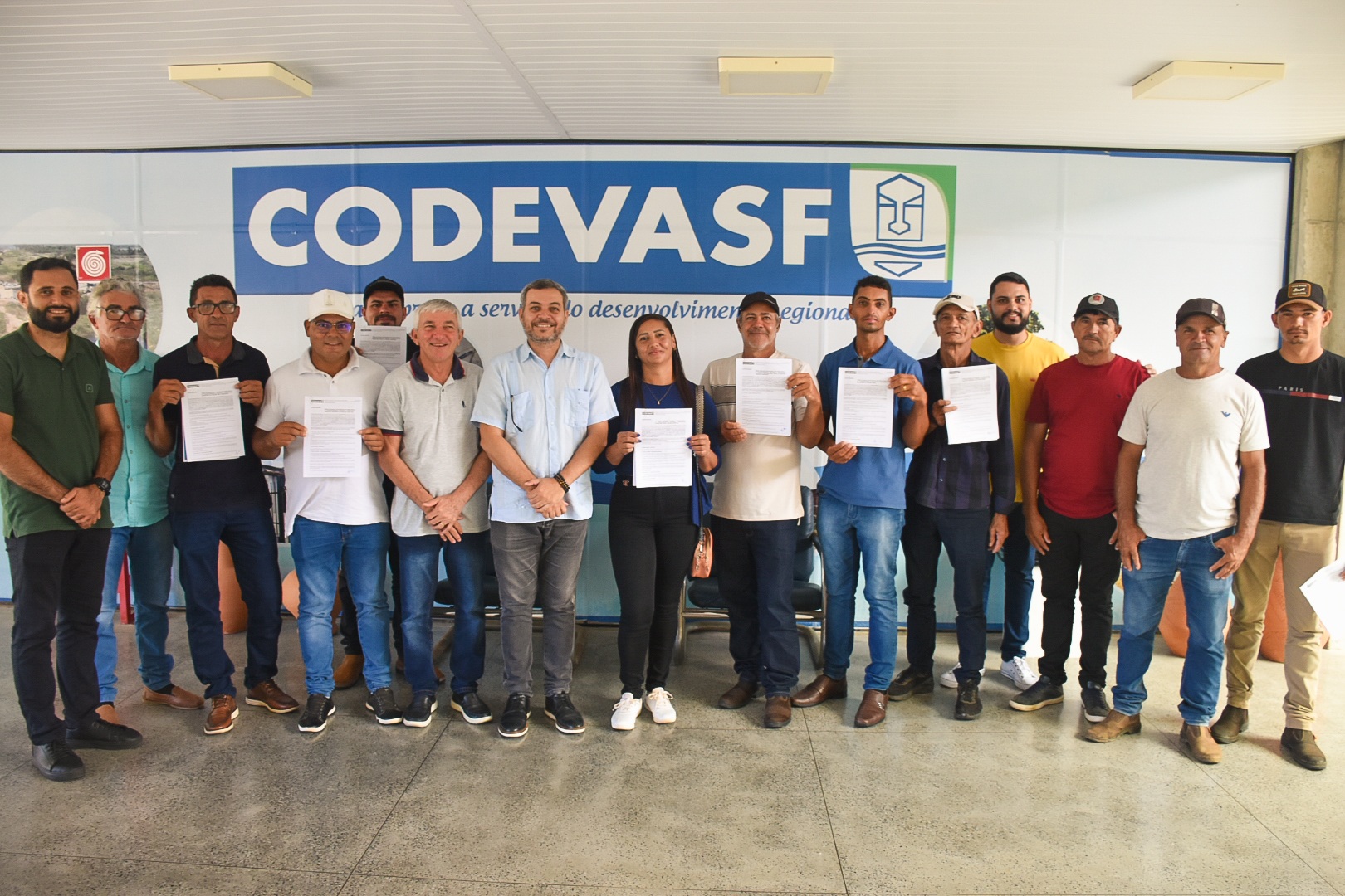 Pessoas posam alinhadas em frente a painel azul e branco, algumas segurando papéis impressos.