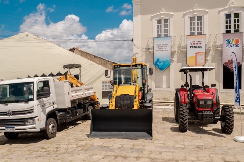 Caminhão, retroescavadeira e trator expostos em evento de entrega de equipamentos.
