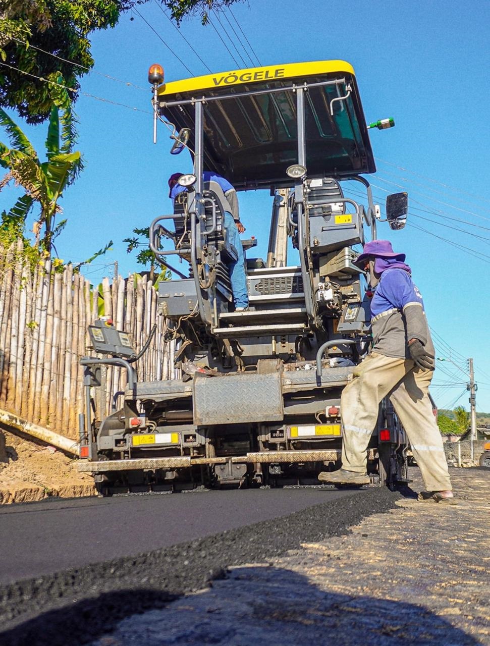 Pavimentadora aplica asfalto em via enquanto dois trabalhadores acompanham o serviço.