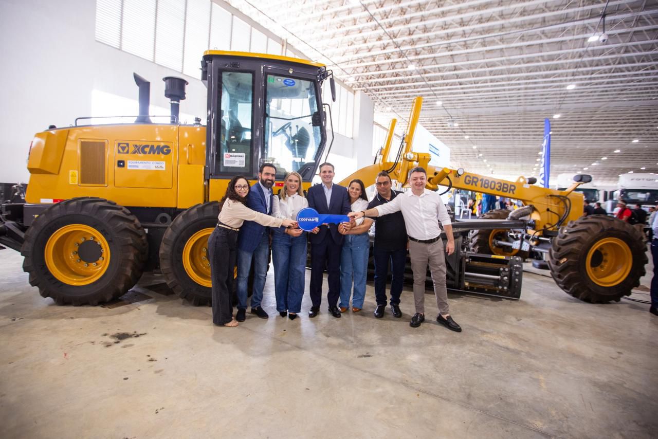 Grupo posa diante de motoniveladora amarela em espaço de exposição coberto