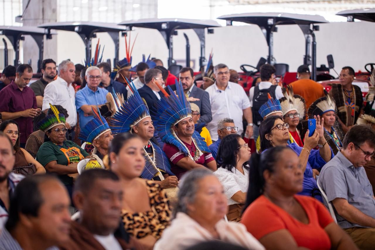 Pessoas assistem a evento; indígenas usam cocares azuis em área coberta