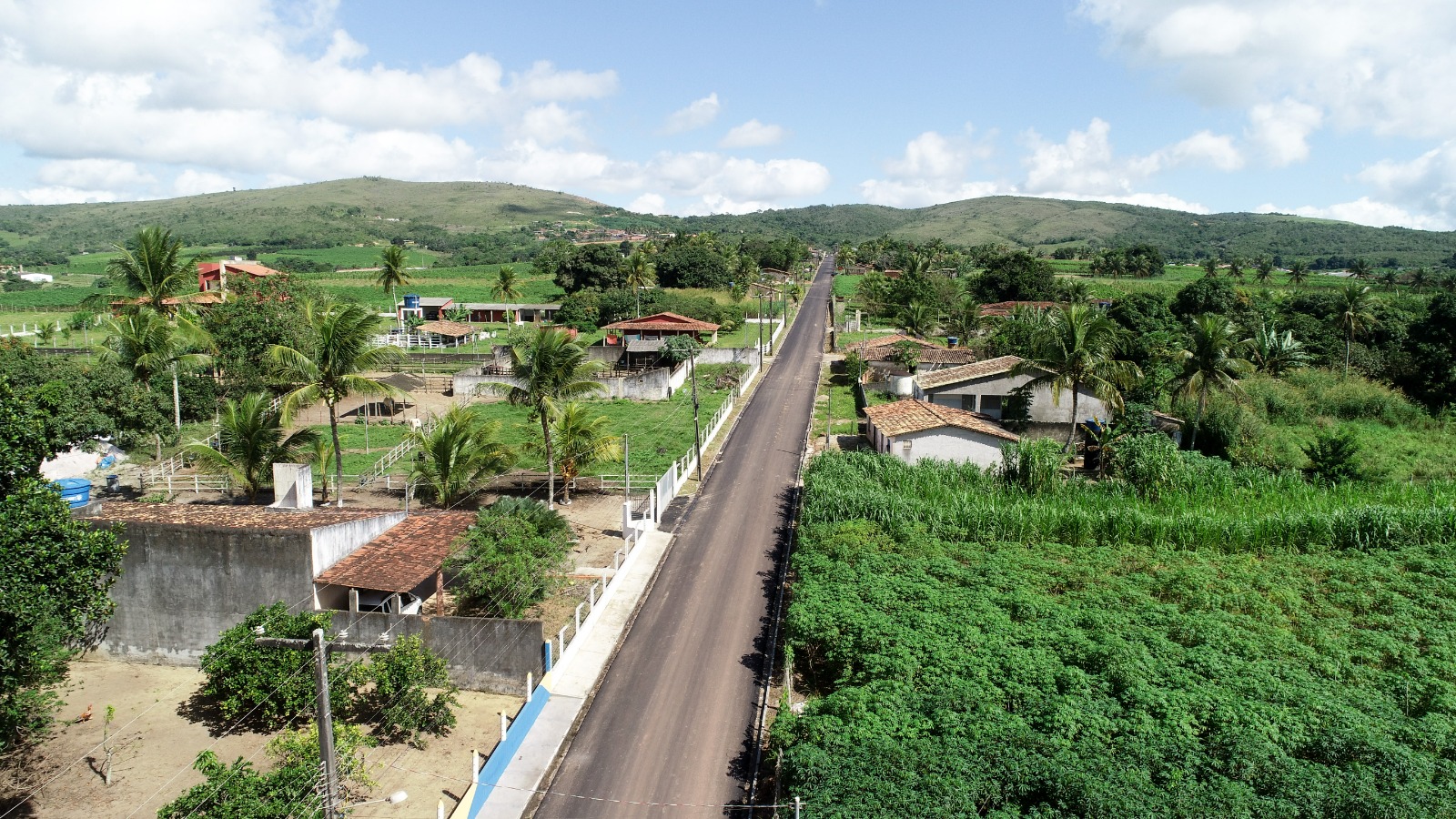 Vista aérea de rua asfaltada cercada por vegetação, casas e colinas ao fundo sob céu com nuvens.