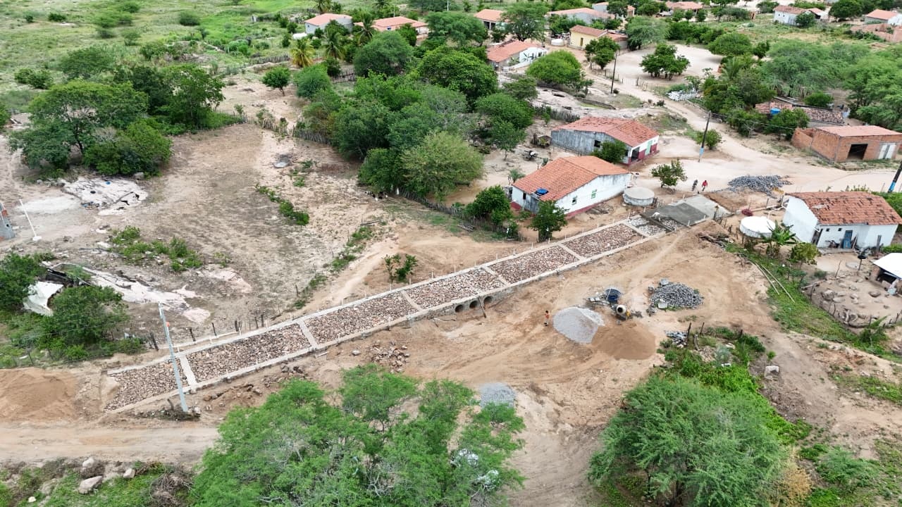 Vista aérea de uma estrada de pedras em obras cortando um terreno de terra com casas e árvores