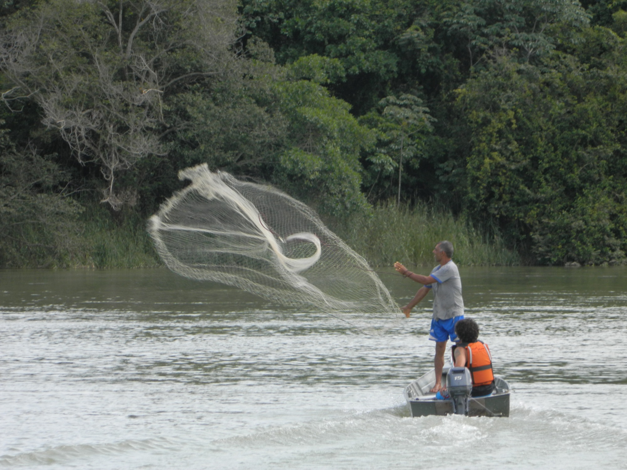 Homem de pé em um barco motorizado joga uma rede de pesca aberta sobre a água de um rio.