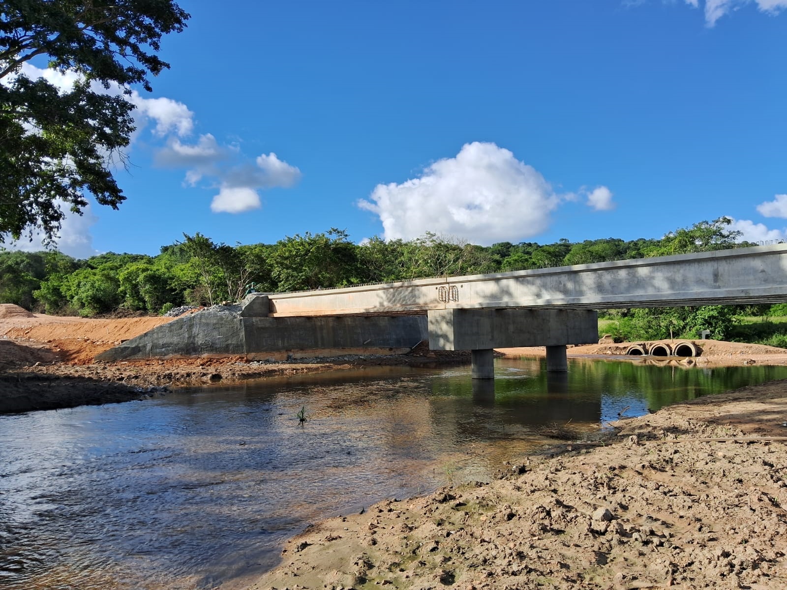 Ponte de concreto sobre rio, com margens de terra e vegetação ao redor.