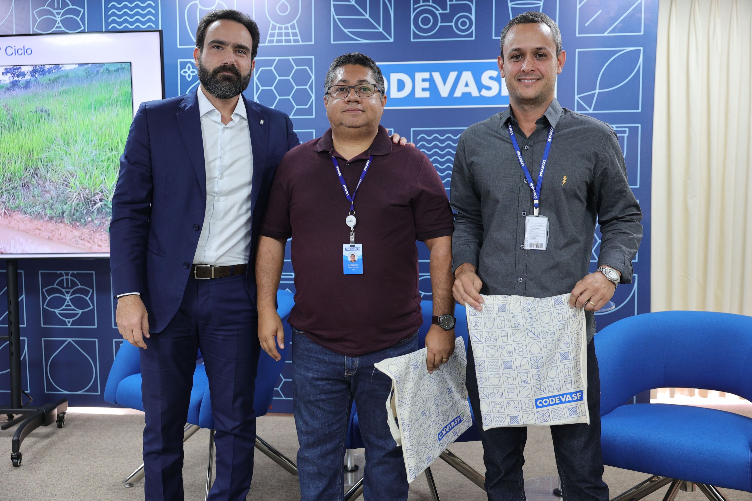 Três homens em pé diante de painel da Codevasf, dois seguram bolsas institucionais.