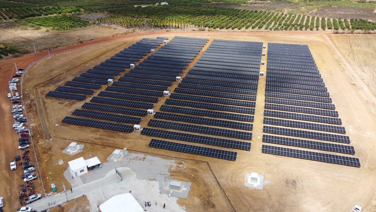 Vista aérea de usina solar com fileiras de painéis fotovoltaicos instalados em terreno cercado.