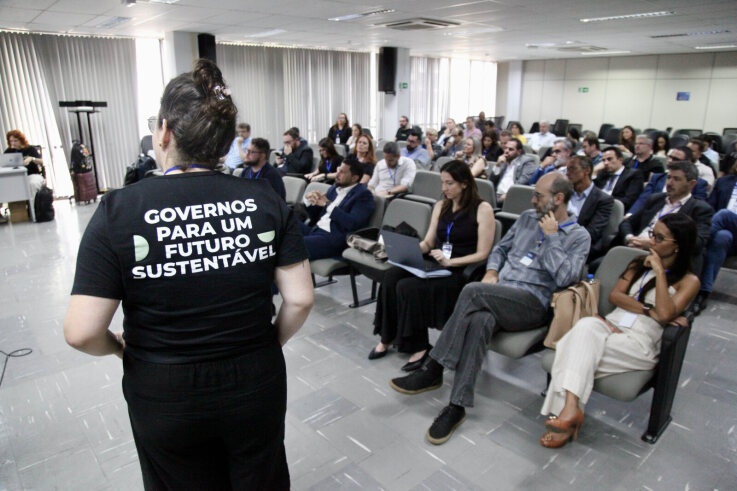 Pessoas sentadas em auditório e mulher com camiseta escrito "Governos para futuro sustentável"