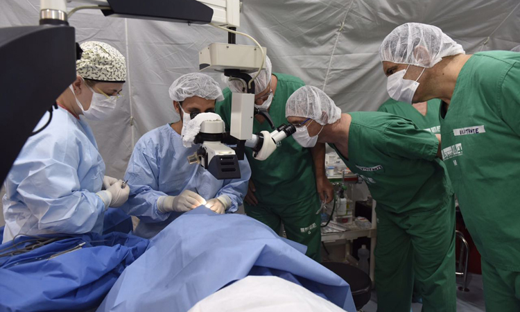 Equipe médica posicionada ao redor da mesa de cirugia realiza procedimento em paciente deitado.