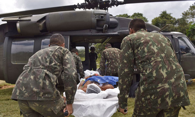 militares carregam uma maca com um paciente deitado sobre ela para um helicóptero pouxa no gramado da aldeia Crispim