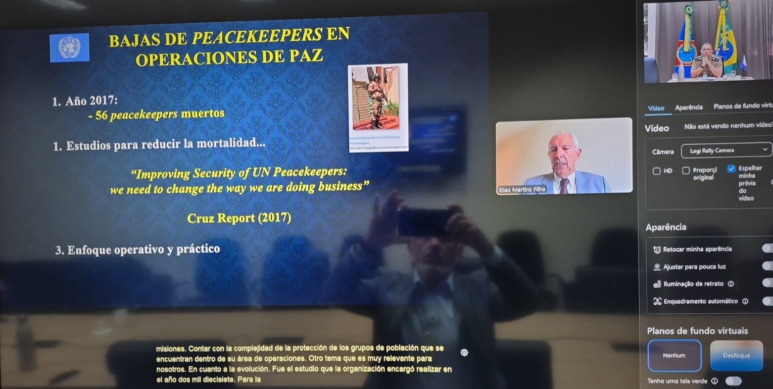 1. Conteúdo do Slide Principal O slide ocupa a maior parte do lado esquerdo da tela, com um fundo azul escuro decorado com marcas d'água do logotipo da ONU.  Título: "BAJAS DE PEACEKEEPERS EN OPERACIONES DE PAZ" (Baixas de mantenedores da paz em operações de paz).  Logotipo: No canto superior esquerdo, vê-se o emblema oficial da Organização das Nações Unidas (ONU).  Tópicos em destaque:  1. Año 2017: Indica que houve 56 peacekeepers mortos naquele ano.  2. Estudios para reducir la mortalidad: Menciona estudos para reduzir a mortalidade.  Citação em destaque: "Improving Security of UN Peacekeepers: we need to change the way we are doing business" (Cruz Report, 2017). Esta frase refere-se ao Relatório Santos Cruz.  3. Enfoque operativo y práctico: (Enfoque operativo e prático).  Imagem interna: Há uma pequena foto de um soldado com uniforme de camuflagem de deserto e capacete azul, em posição de guarda.  2. Participantes da Reunião Dois quadros de vídeo são visíveis no lado direito:  Palestrante Principal: Um homem de cabelos brancos, usando terno azul e gravata bordô, identificado pela legenda como Elias Martins Filho (ex-Comandante da Força da MONUSCO).  Participante Adicional: No canto superior direito, um oficial militar fardado aparece sentado em uma mesa com bandeiras ao fundo (incluindo a bandeira do Brasil).  3. Interface do Software e Legendas Configurações: No lado direito, há um painel de controle de vídeo com opções como "Retocar minha aparência", "Ajustar para pouca luz" e "Planos de fundo virtuais".  Legendas em tempo real: Na parte inferior da tela, aparecem legendas automáticas em espanhol que transcrevem a fala sobre a complexidade da proteção de grupos populacionais e a evolução dos estudos encomendados pela organização em 2017.  Reflexo: É possível notar o reflexo da pessoa que tirou a foto da tela, segurando um smartphone em frente ao monitor.  4. Contexto Técnico A imagem registra um momento de compartilhamento de conhecimento sobre segurança militar internacional, especificamente focado no Relatório Cruz de 2017, que foi um marco na mudança de estratégia das missões de paz da ONU para reduzir fatalidades de "capacetes azuis".