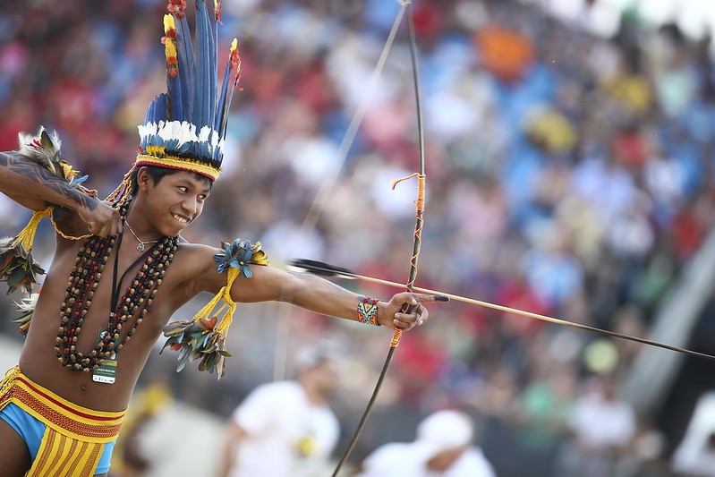 Jovem indígena com pintura corporal e cocar atira flecha durante evento cultural