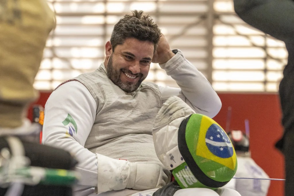 Com a mão nos cabelos, atleta sorri enquanto segura o capacete e a espada.