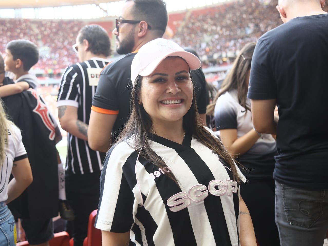 Torcedora com camisa do Corinthians e boné rosa, cabelos lisos e loiros sorri para a foto.