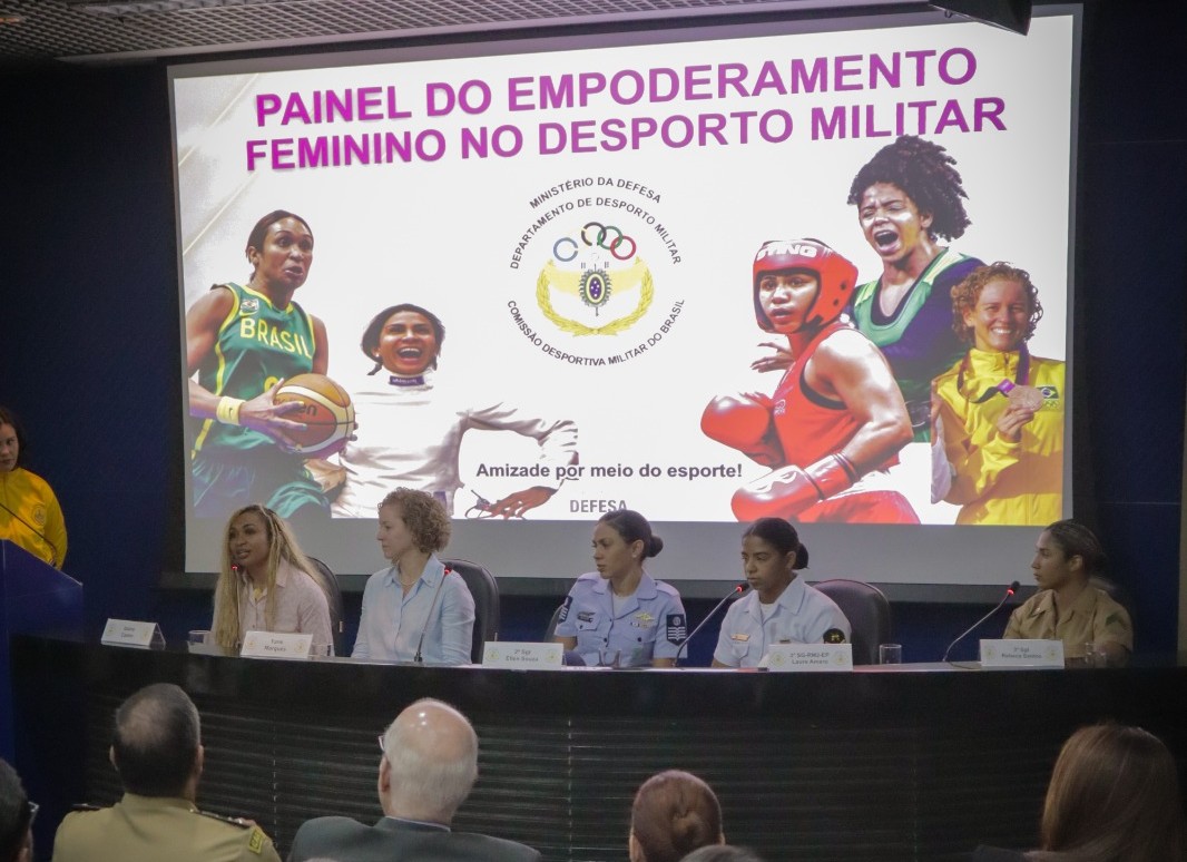 Mesa com participação das atletas e da secretária Iziane Marques. Ao fundo há um letreiro dizendo mu