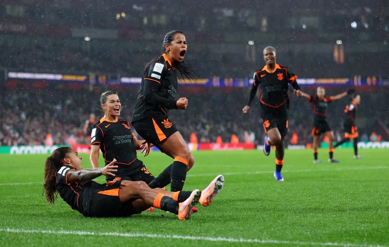 Ao centro jogadora comemora gritando. Duas caídas no chão também sorriem e gritam, enquanto outra se aproxima correndo.
