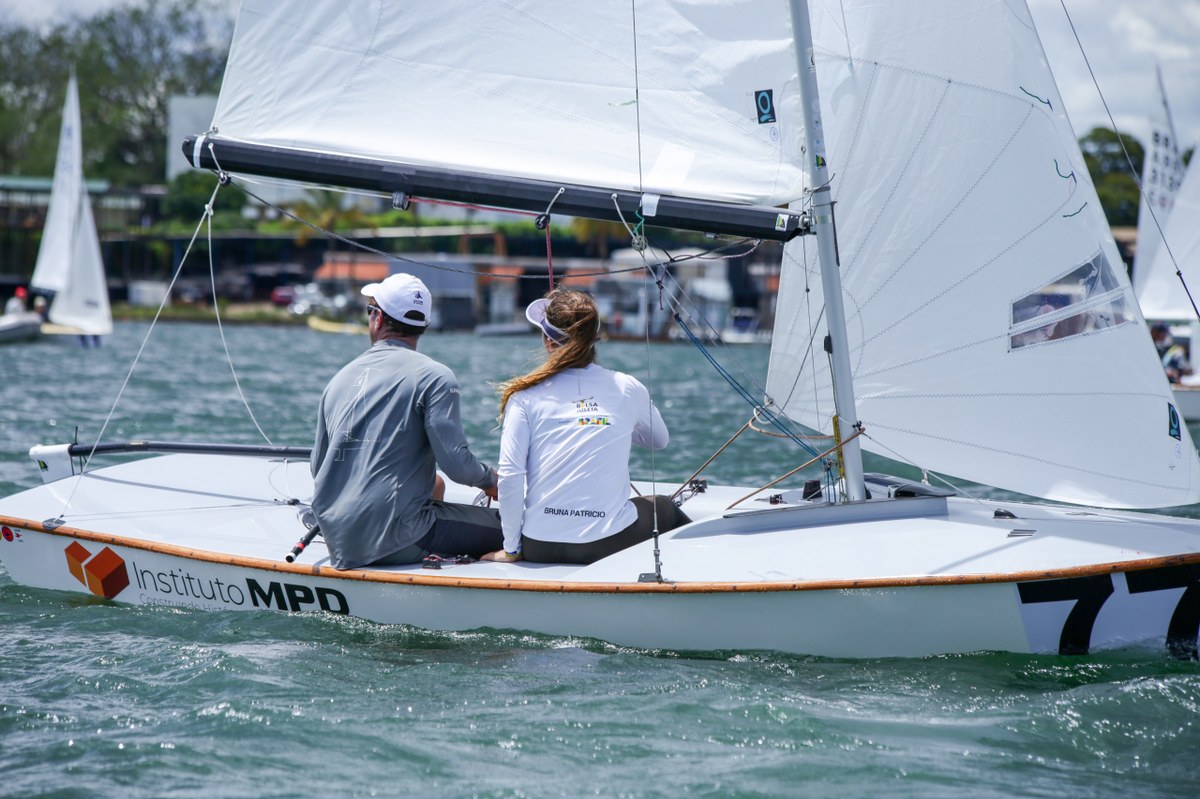 Dupla de velejadores de costas. Um homem de camisa de manga longa cinza e uma mulher de camisa de manga longa branca.