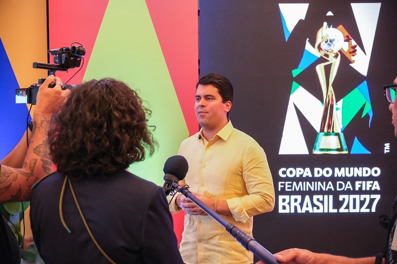 Ministro do esporte, com camisa amarela, dando entrevista a uma jornalista que está de costas.