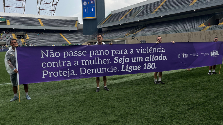 Faixa lilás no campo com os dizeres: Não passe pano para a violência contra a mulher. Seja um aliado. Proteja. Denuncie. Ligue 80.