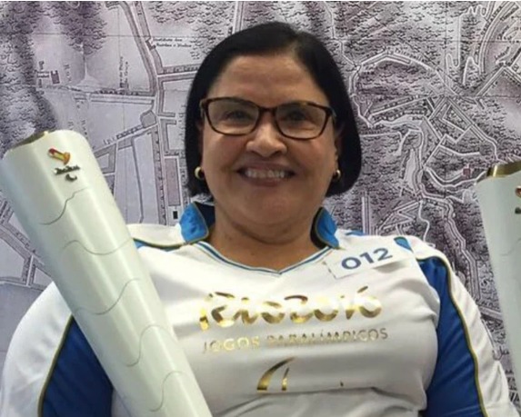 mulher de cor parda, cabelos lisos e curtos usando óculos e brincos, com sorriso no rosto, vestindo uniforme de competição e segurando uma tocha olímpica. 