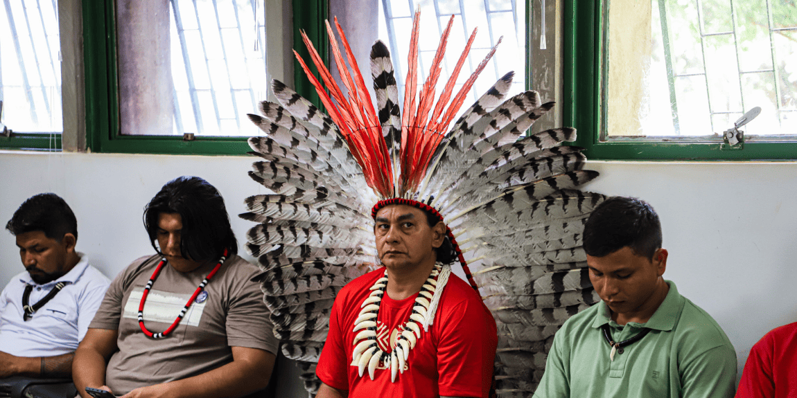 Liderança indígena sentada em sala de reunião, usando grande cocar de penas vermelhas e brancas em formato de leque, colar tradicional e camisa vermelha, ao lado de outros participantes indígenas e não indígenas, com rostos desfocados, em ambiente interno com janelas ao fundo.