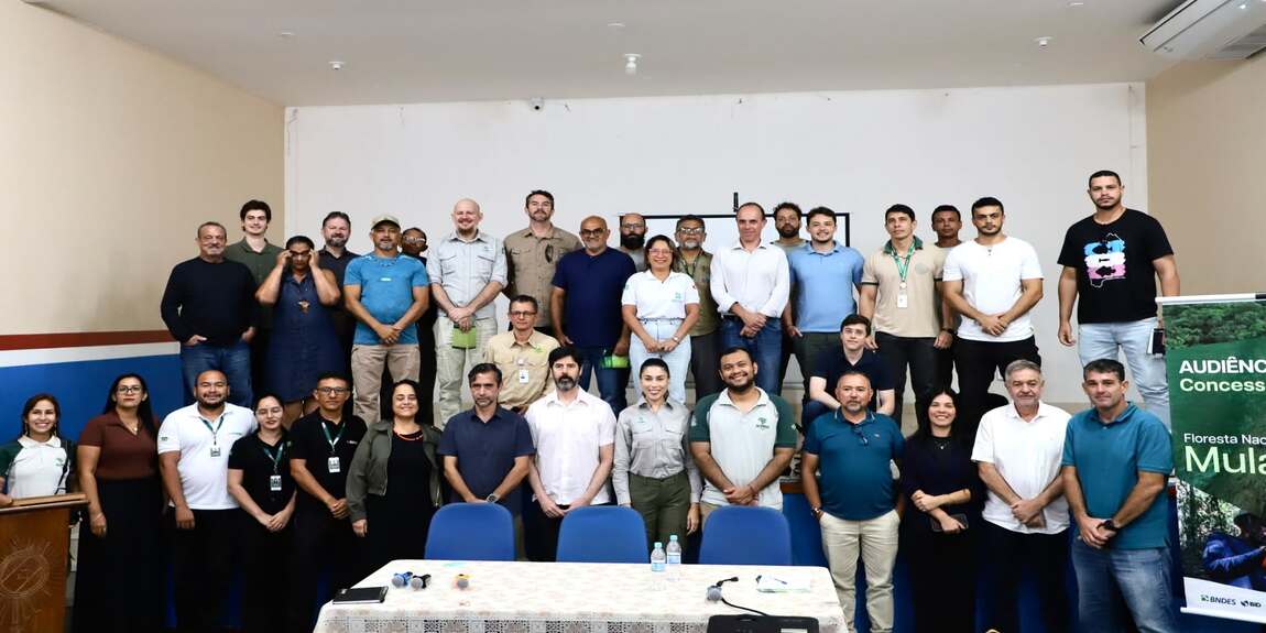 Fotografia de um grupo com cerca de 35 pessoas reunidas em um auditório. Os participantes estão organizados em três fileiras, posando para a foto em frente a uma parede branca. À direita, há um banner da audiência pública sobre a concessão florestal da Floresta Nacional de Mulata. À frente do grupo, uma mesa com cadeiras, microfones e garrafas de água.