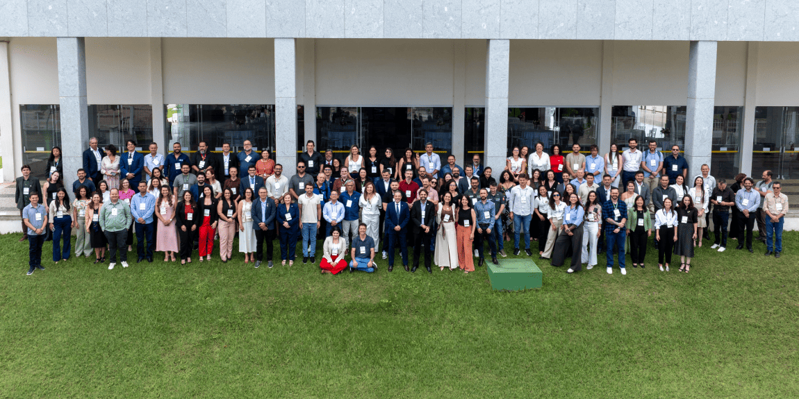 Foto de grupo ao ar livre, com muitas pessoas reunidas em frente a um edifício institucional.