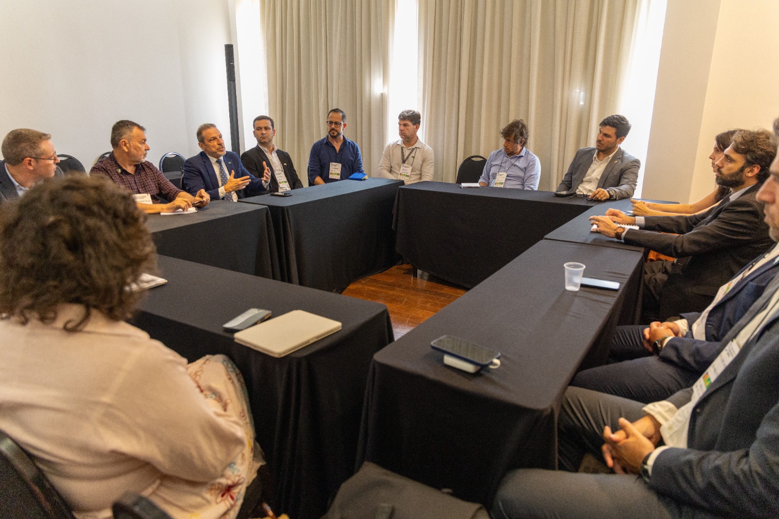 Foto de reunião com pessoas sentadas ao redor de mesas em formato de U, em ambiente interno.