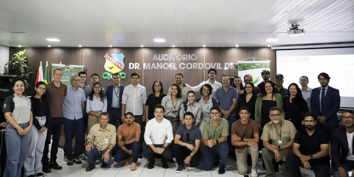 Fotografia colorida de um grupo de aproximadamente 23 pessoas reunidas para uma foto coletiva em um auditório. Parte do grupo está em pé, ao fundo, e outra parte está sentada ou agachada, à frente. As pessoas vestem roupas variadas, predominantemente em tons neutros, azul, verde, preto e branco. Ao fundo, há um painel de madeira com a identificação do Auditório Dr. Manoel Correia Diniz, além de logomarcas institucionais. À esquerda, aparecem as bandeiras do Brasil e de outro estado, e um painel relacionado a edital de concessão e restauração florestal. Ao centro e à direita, há banners institucionais do Serviço Florestal Brasileiro. No lado direito da imagem, vê-se uma tela de projeção. O grupo posa sorrindo e olhando para a câmera.