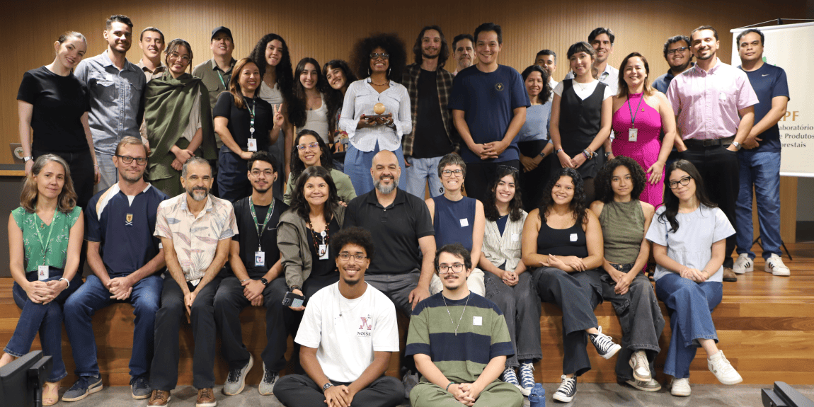 Foto de grupo com cerca de 30 pessoas reunidas em um auditório, organizadas em três fileiras. Ao fundo, há um painel de madeira e um banner institucional.
