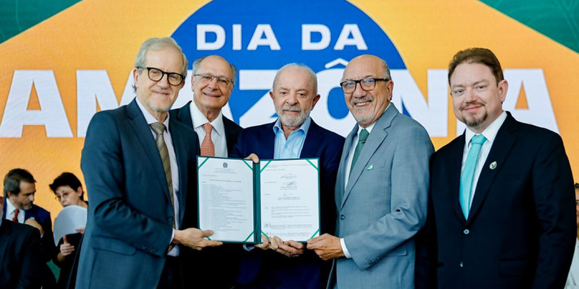 Cinco homens sorrindo seguram um documento assinado, diante de um painel com a palavra “Amazônia” ao fundo.