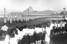 Fotografia em preto e branco de soldados brasileiros marchando em formação em uma avenida, observados por mulheres e meninas, com montanhas ao fundo.