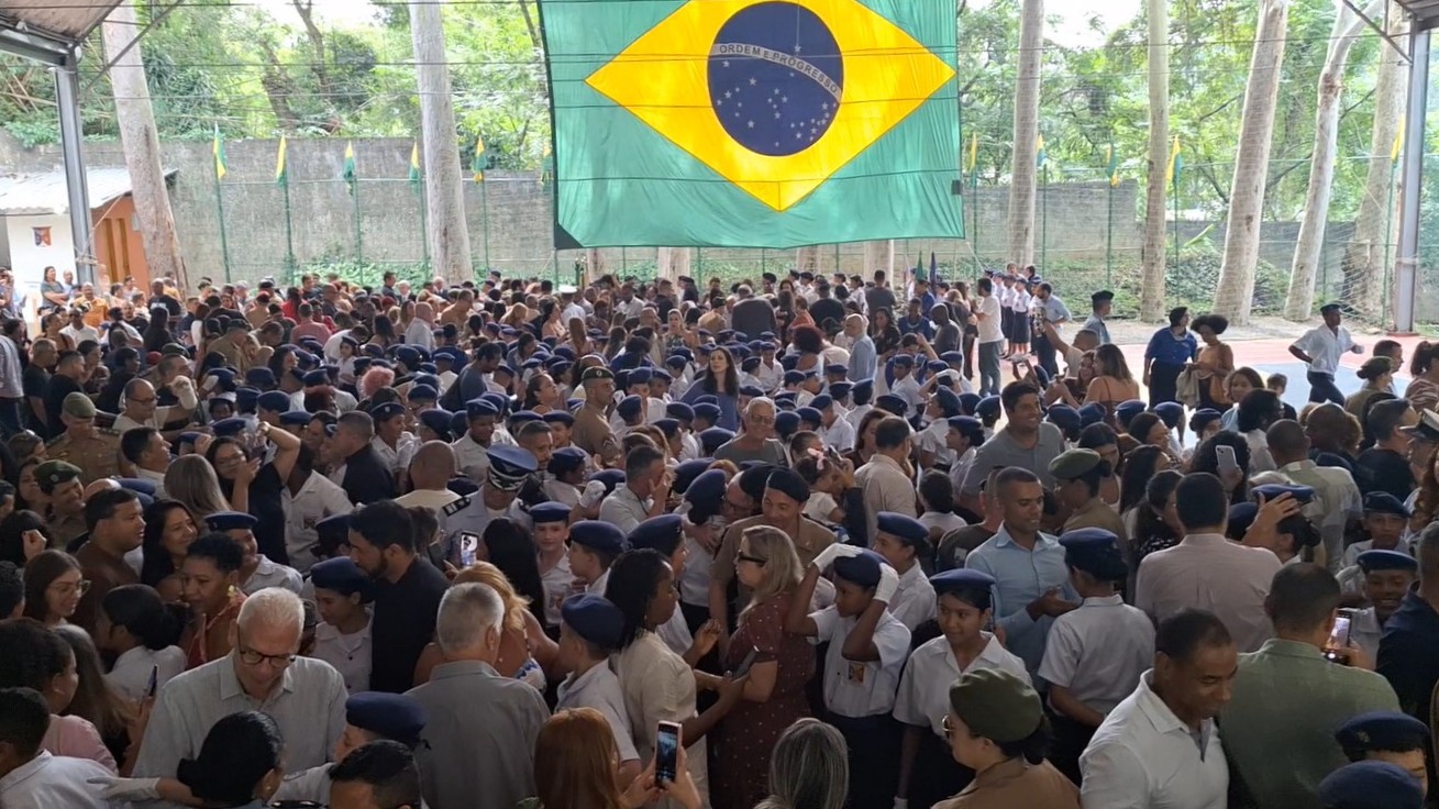 Público e estudantes reunidos sob bandeira do Brasil em cerimônia escolar militar.