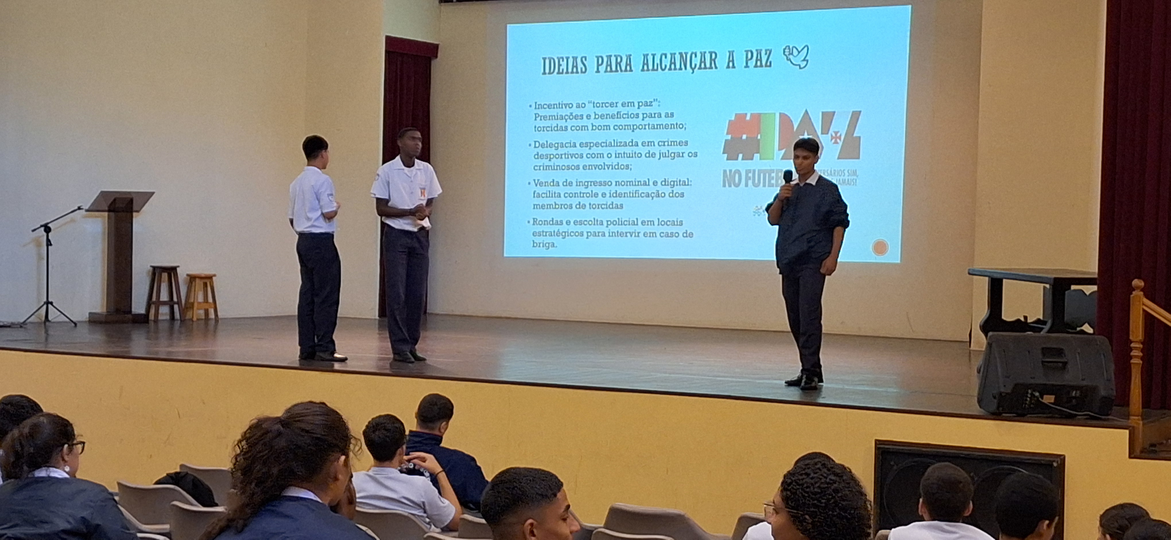 Três alunos apresentam trabalho no palco com slide projetado sobre ideias para alcançar a paz.