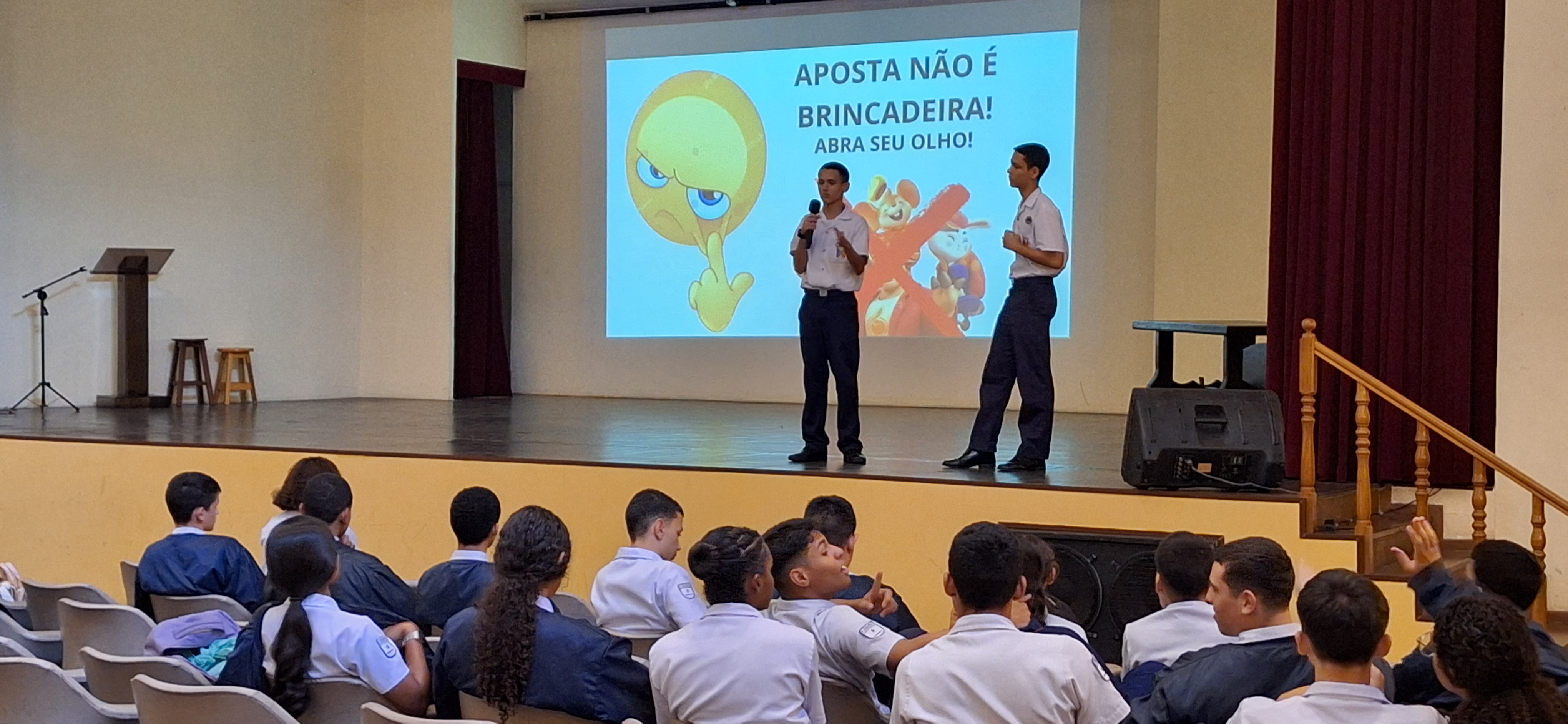 Dois alunos apresentam no palco com slide escrito “Aposta não é brincadeira”, enquanto estudantes assistem sentados.