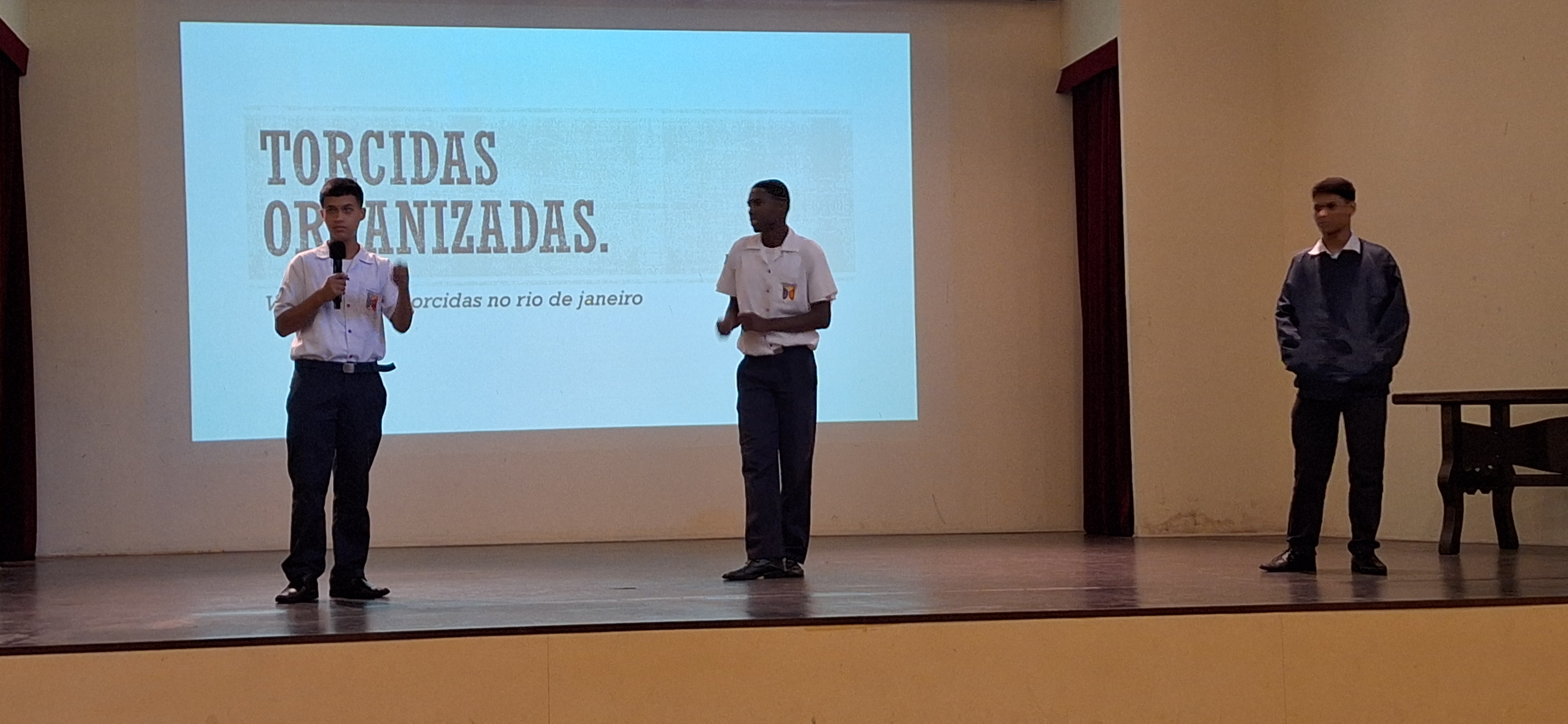 Três alunos apresentam trabalho no palco com slide projetado sobre ideias para alcançar a paz.