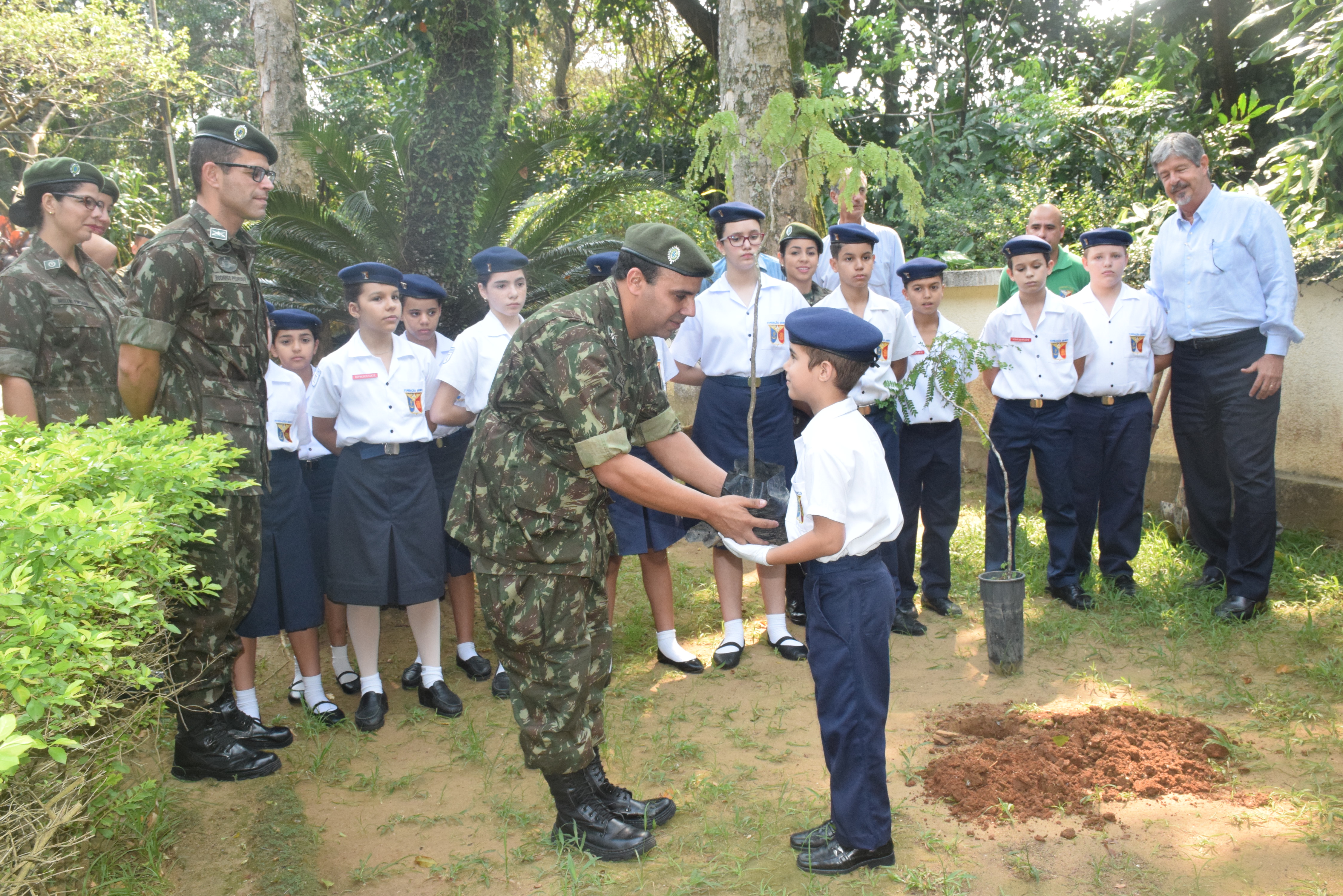 Militar entrega uma muda de árvore a um aluno fardado, enquanto colegas e adultos observam a ação em área verde.