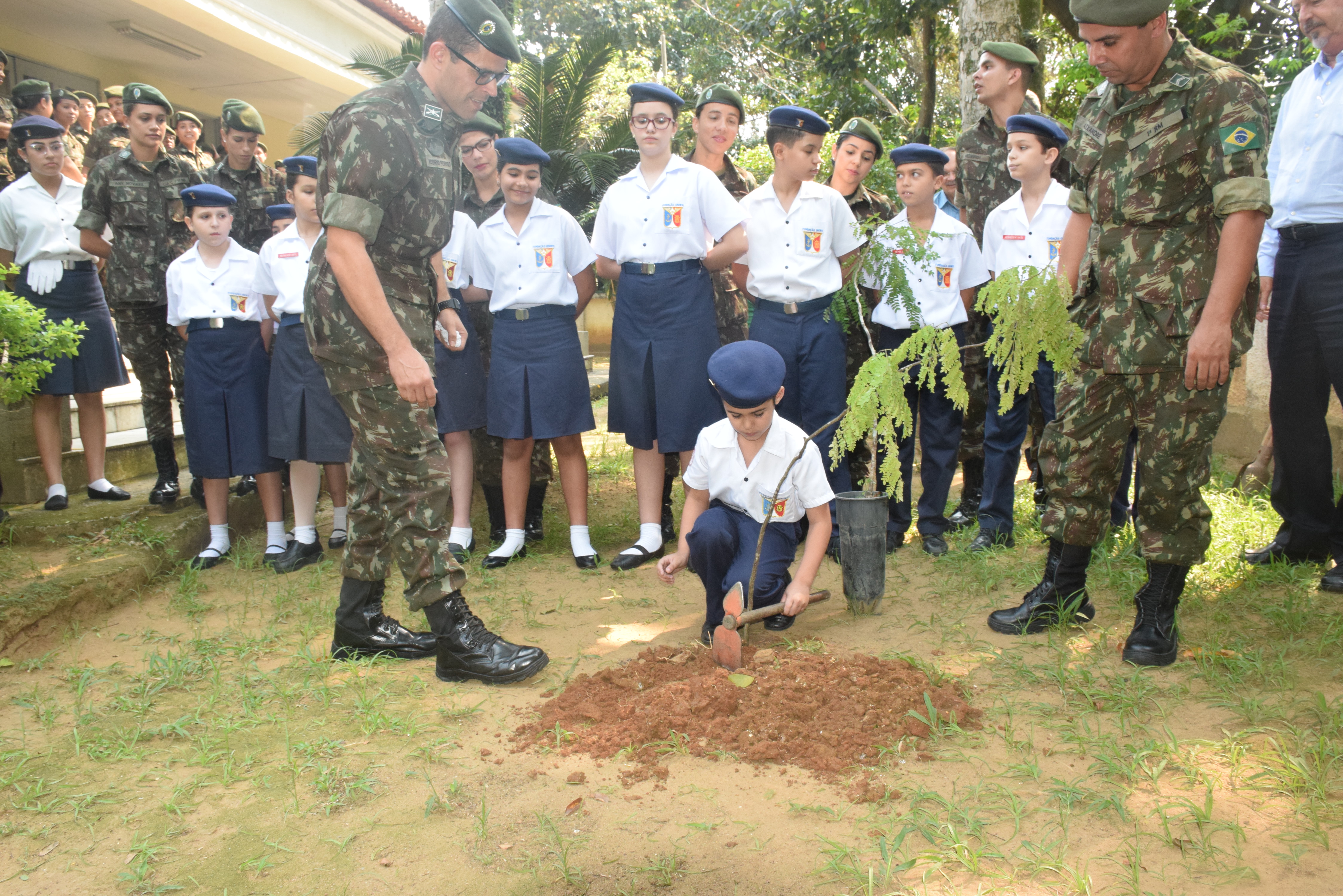 Aluno fardado planta uma muda de árvore no solo, com acompanhamento de militares e outros estudantes durante atividade ambiental