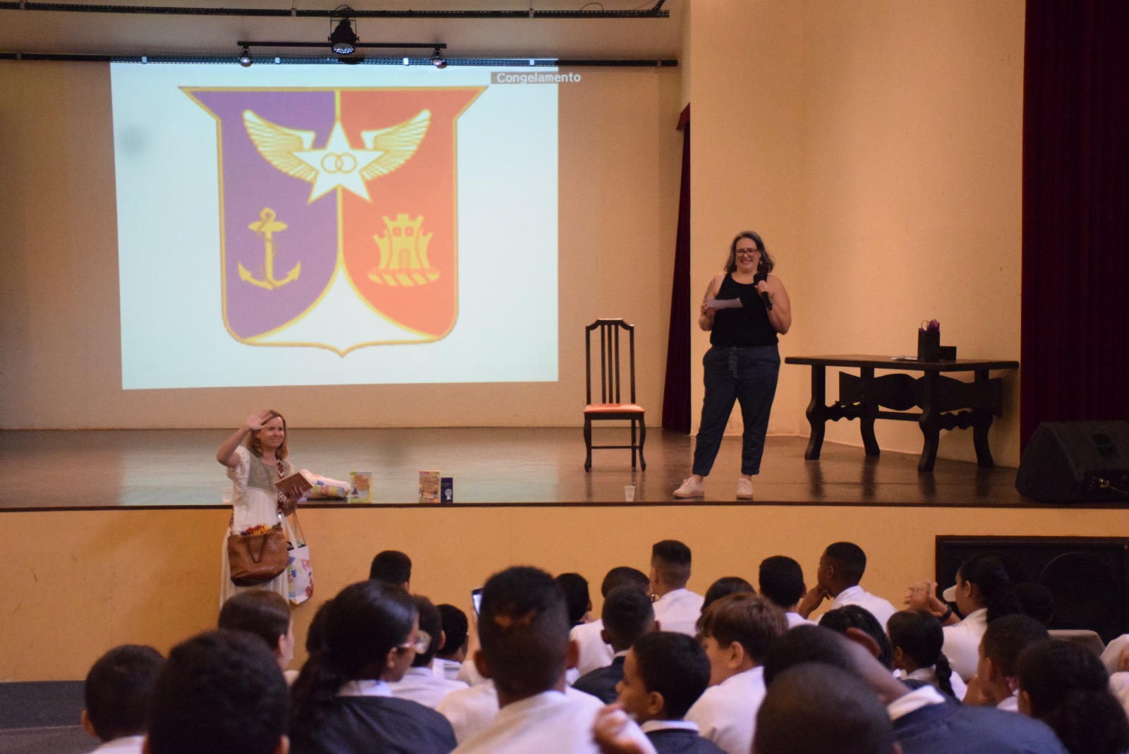 Mulher em pé no palco falando ao microfone para alunos sentados no auditório, com brasão projetado ao fundo.