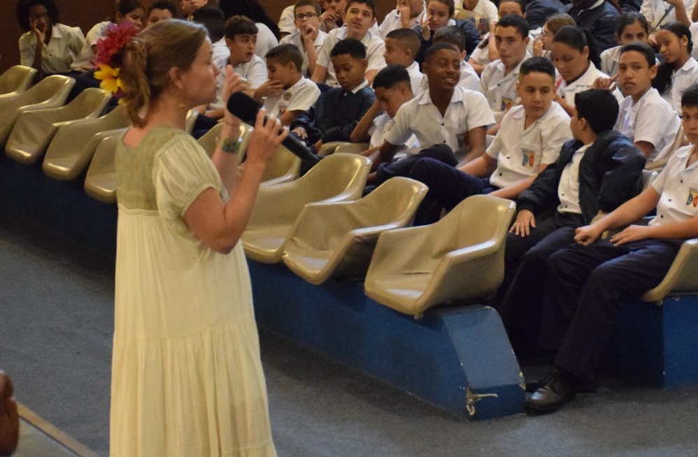 Mulher falando ao microfone para estudantes sentados em cadeiras de auditório durante uma apresentação.