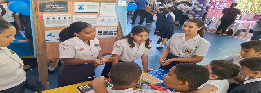 Estudantes reunidos em torno de um estande científico com cartazes e maquetes, durante evento escolar.