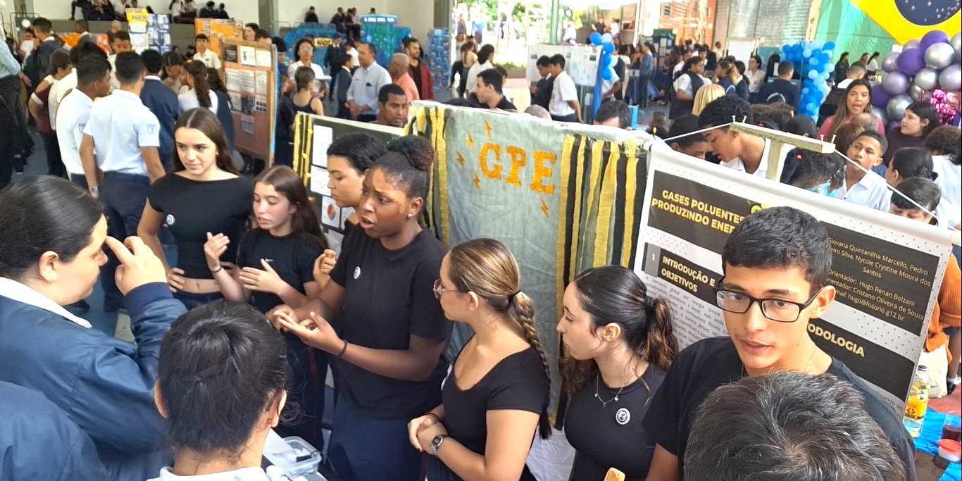 ssor conversa com grupo de estudantes que explicam um experimento em um estande de feira científica.