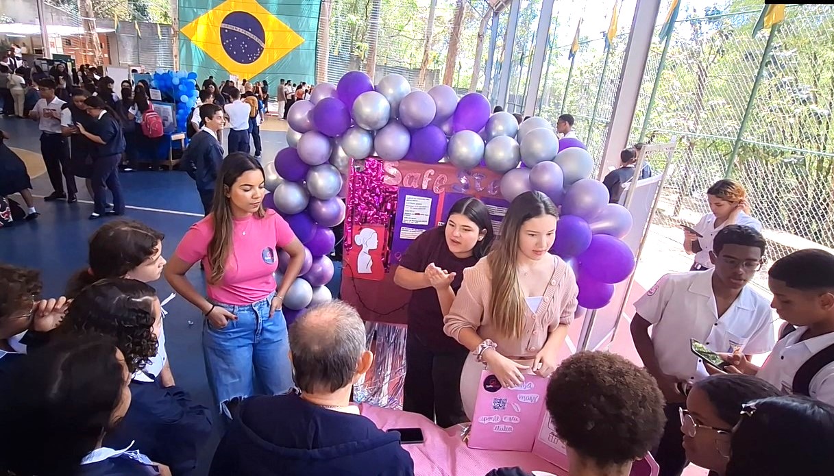 Estande decorado com balões roxos e prateados, alunas apresentam projeto para visitantes em uma feira escolar.
