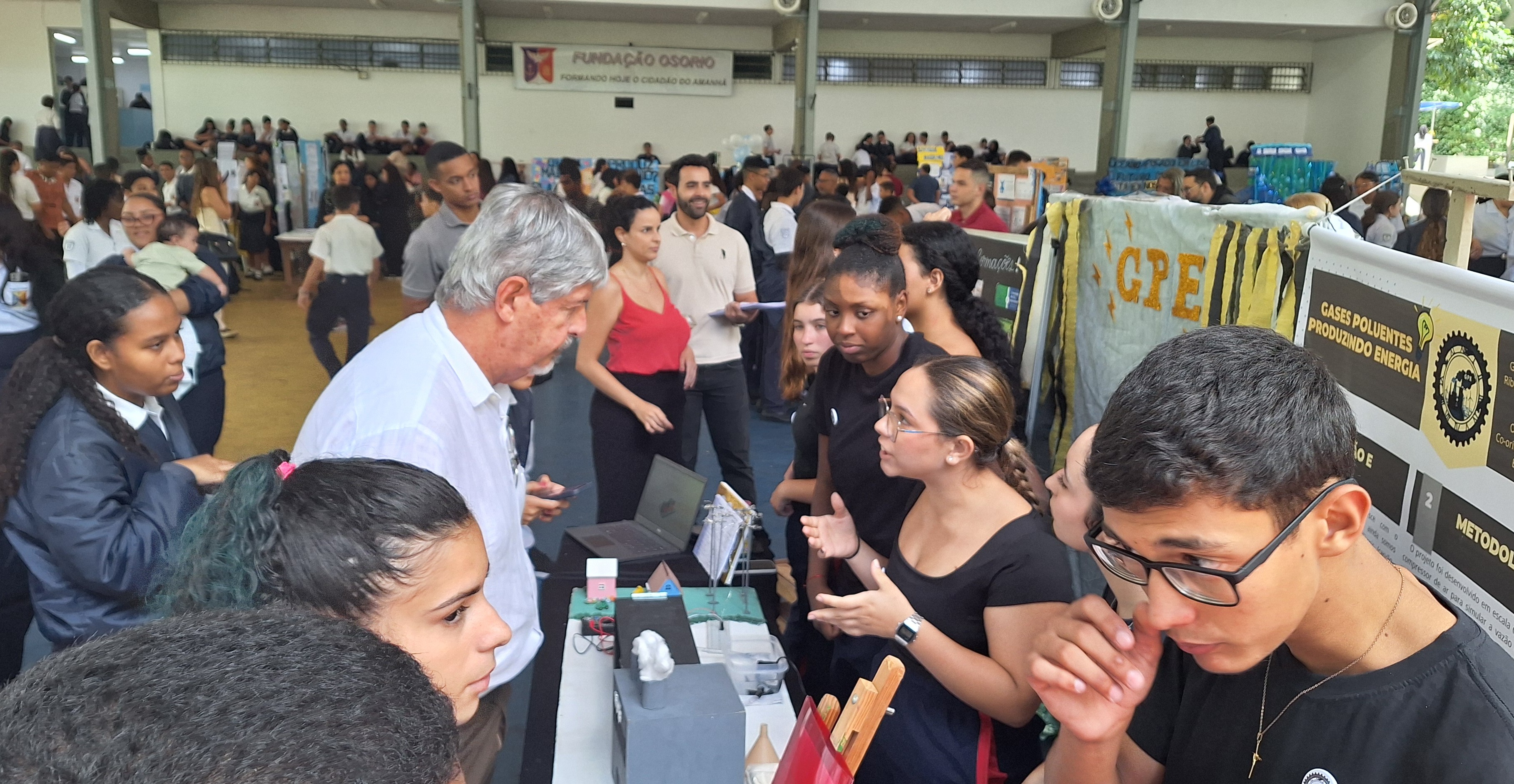 ssor conversa com grupo de estudantes que explicam um experimento em um estande de feira científica.