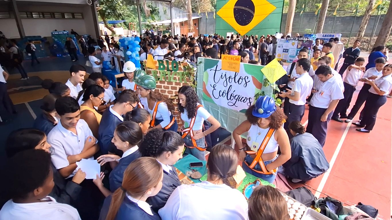Vista aérea de estande com o tema “Tijolos ecológicos”, estudantes interagem com visitantes durante feira escolar.