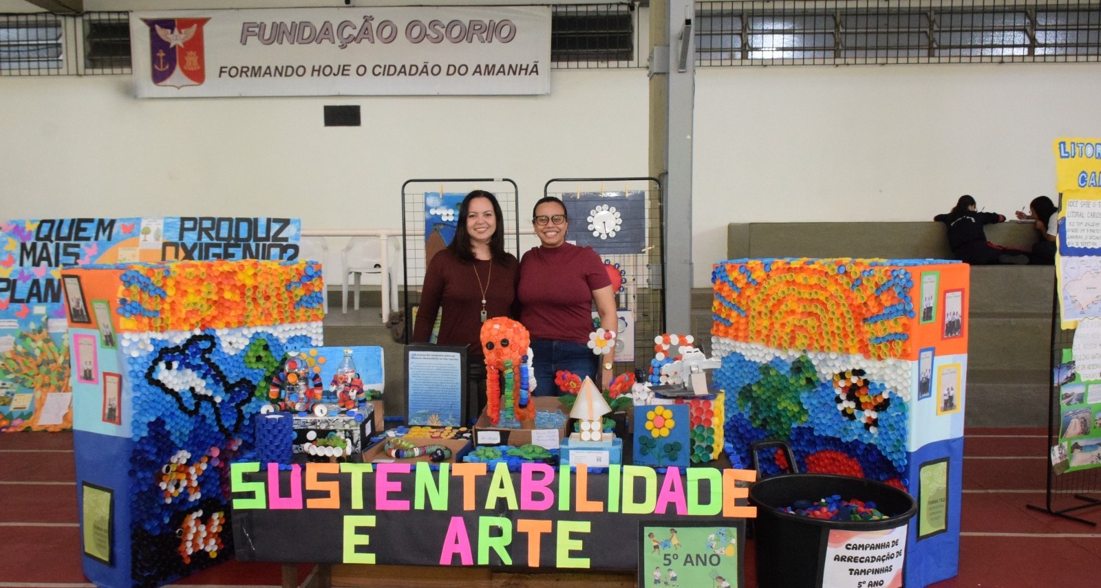 Professoras posam sorrindo atrás de um estande com trabalhos feitos de materiais recicláveis, com o tema “Sustentabilidade e Arte”.