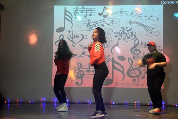 Grupo de jovens dançando no palco com fundo de notas musicais projetadas.